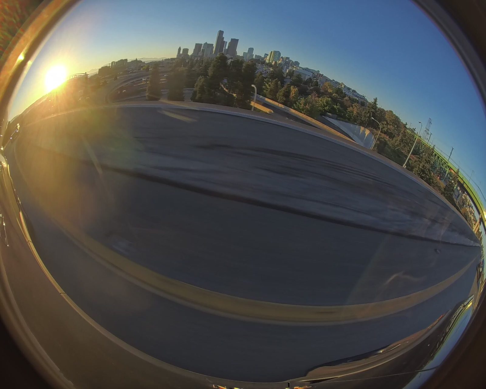 A side-camera fisheye frame on the I-5/I-90 interchange curve at sunset: the low sun flares at the left edge, and the downtown Seattle skyline stands above the ramps and trees.